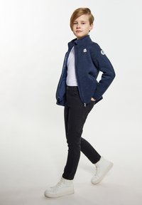 Tamsiai mėlyna užtraukiama striukė su tekstūruotu paviršiumi, balta marškinėliai, juodos aptemptos kelnės ir balti aukšti sportbatiai su raišteliais.