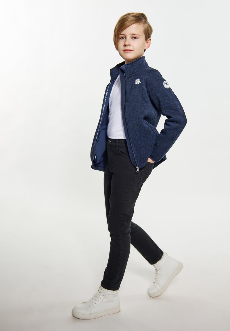 Tamsiai mėlyna užtraukiama striukė su tekstūruotu paviršiumi, balta marškinėliai, juodos aptemptos kelnės ir balti aukšti sportbatiai su raišteliais.