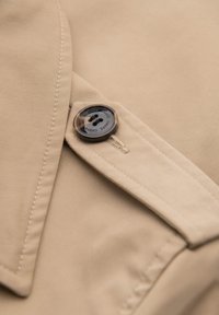 Gros plan sur un tissu beige avec un bouton marron fixé à une sangle cousue sur un vêtement, mettant en valeur la texture et les détails de la couture.