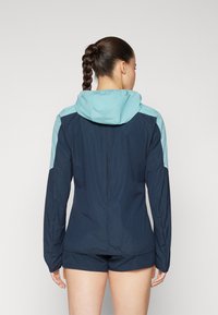 Marineblå lett jakke med en teal aksent på hetten og skuldrene. Har lange ermer og en slank, tettsittende design.
