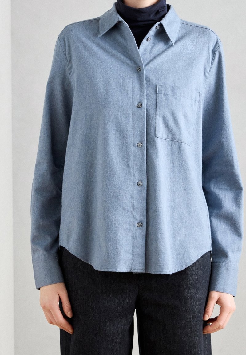 Personne portant une chemise bleu clair à boutons par-dessus un col roulé noir, associée à un pantalon foncé, vue du cou aux hanches.