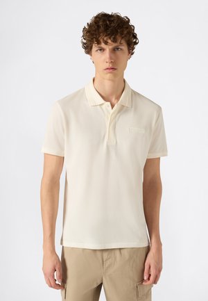 Joven con cabello rizado que lleva una camiseta tipo polo crema y pantalones cargo beige, de pie contra un fondo blanco liso.