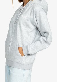 Femme portant un sweat à capuche gris clair avec fermeture éclair et poches, ainsi qu'un jean bleu clair, debout de profil avec une main dans la poche.