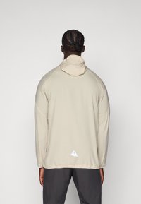 Beige hooded jacket with long sleeves, featuring a drawstring, textured fabric, and a white triangular logo at the lower hem.