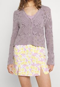 Lila öppen stickad cardigan med vågig nederkant, i kombination med en vit kjol med ett gult och rosa blommönster samt enkelsidig slits.