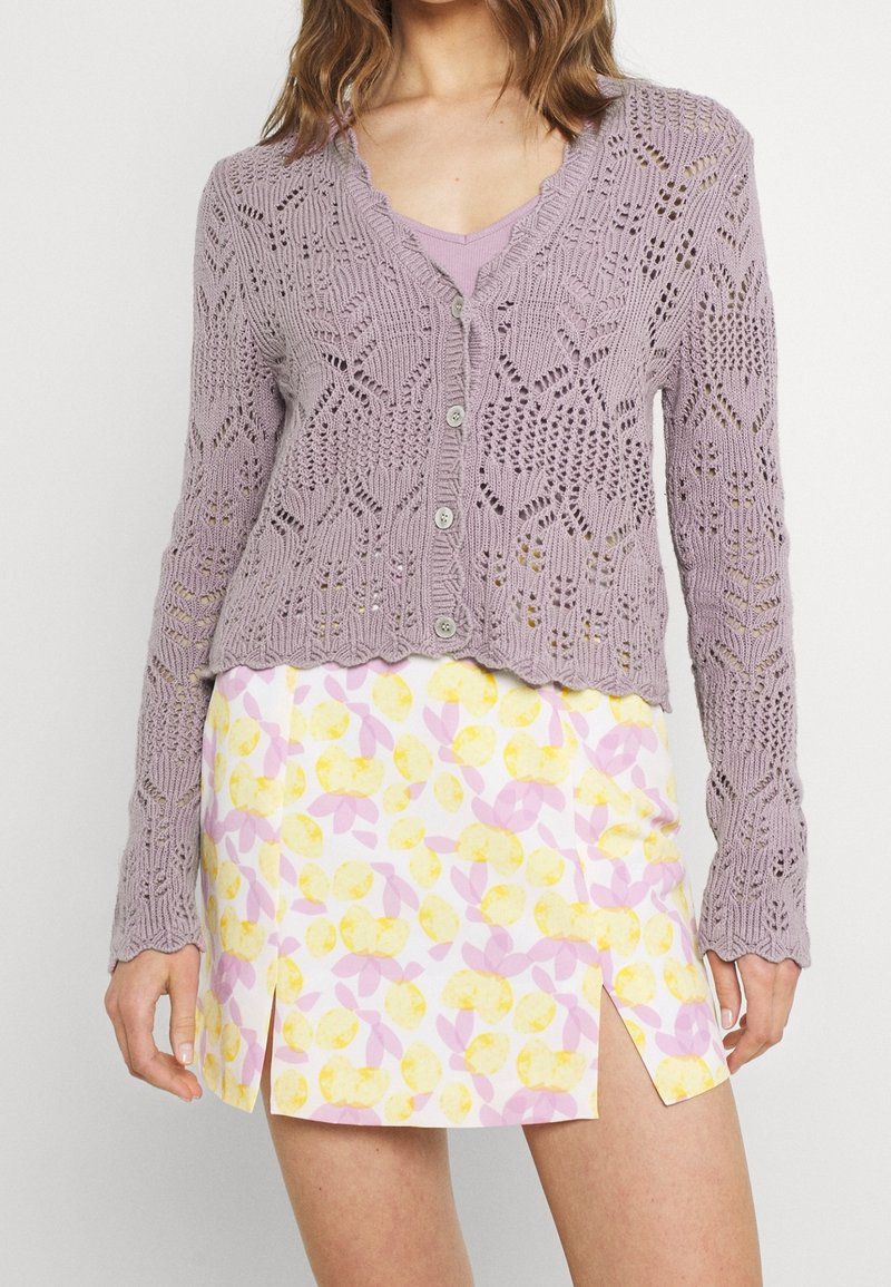 Lila öppen stickad cardigan med vågig nederkant, i kombination med en vit kjol med ett gult och rosa blommönster samt enkelsidig slits.