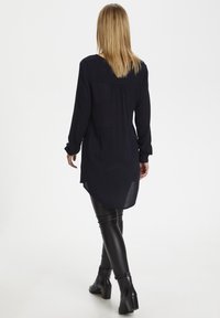 Frau, die davonläuft, trägt eine lange schwarze Bluse, schwarze Lederleggings und schwarze Stiefeletten vor einem schlichten weißen Hintergrund.