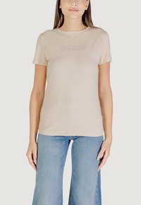 T-shirt beige a maniche corte con collo rotondo e testo "GUESS" in finitura glitterata. Indossato con jeans a vita alta blu chiaro.