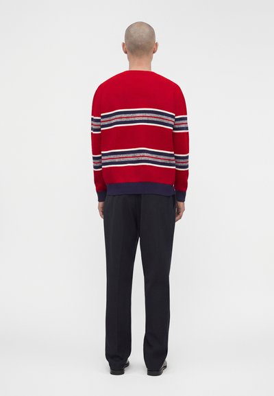 Homme au crâne rasé portant un pull rouge avec des rayures horizontales blanches, bleues et grises, associé à un pantalon noir, debout de dos.