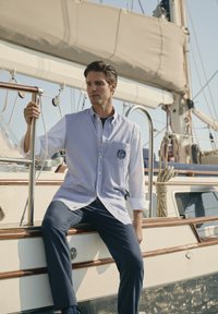 Uomo in camicia a righe e pantaloni blu seduto sulla ringhiera di una barca a vela, mentre tiene un corrimano di metallo, con corde e vele sullo sfondo.