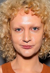 Krøllete blondt hår, lys hud, blå øyne, iført en brun topp. Jevn hudtekstur, nøytral sminke, symmetrisk ansiktsformer, minimale øyenbryn.