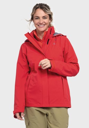Giacca impermeabile rossa con cappuccio, polsini regolabili, cerniera frontale e tasche laterali. Indossata sopra una maglietta arancione, abbinata a pantaloni beige.