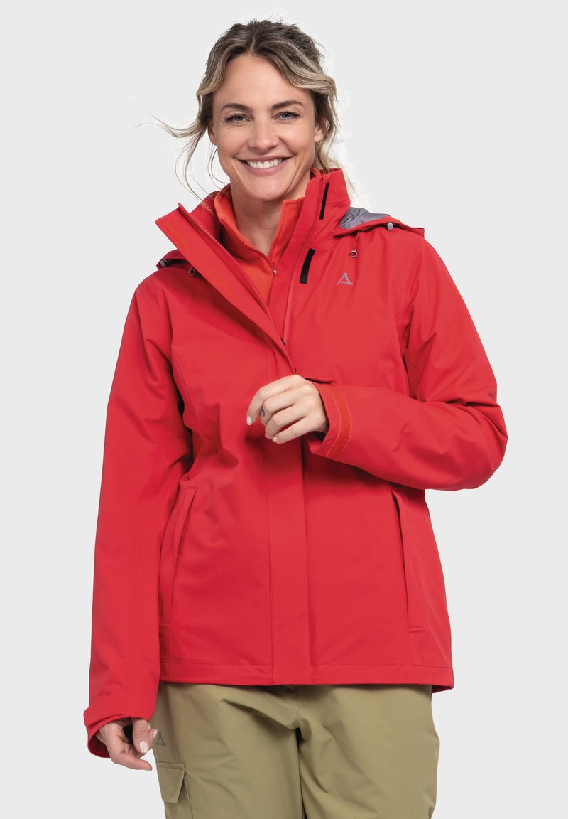 Giacca impermeabile rossa con cappuccio, polsini regolabili, cerniera frontale e tasche laterali. Indossata sopra una maglietta arancione, abbinata a pantaloni beige.
