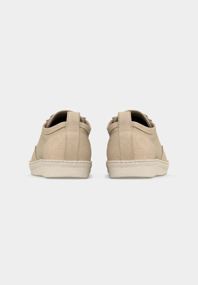 Šviesiai smėlio spalvos laisvalaikio sportbačiai su zomšine kulno dalimi ir tekstūruota balta guminė pado dalimi, parodyti iš nugaros ant vienalytės šviesiai pilkos spalvos fono.