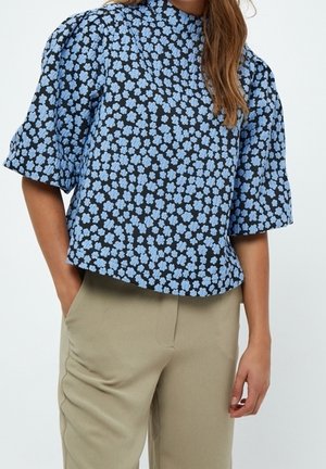 Haut à motifs floraux bleus avec manches courtes bouffantes, col haut, sur fond noir, associé à un pantalon beige clair.