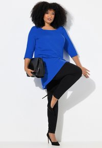 Blå långärmad topp med rundad halsringning, matchad med svarta leggings och högklackade skor. Modellen håller en svart kuvertväska.