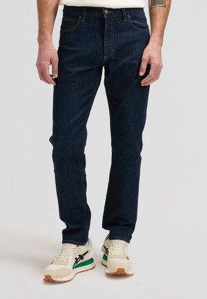 Mężczyzna ubrany w ciemnoniebieskie dżinsy i białe sneakersy z czarnymi i zielonymi akcentami, stojący na tle jednolitego szarego tła.