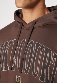 Sweat à capuche marron avec un grand motif texte graphique. Le tissu semble doux avec des coutures texturées et des cordons de serrage.