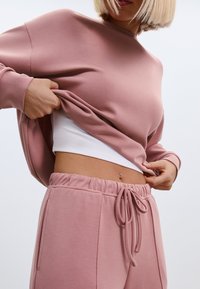 Roze dukserica s širokim rukavima, teksturirane tkanine i okruglim izrezom, u kombinaciji s odgovarajućim ružičastim trenirkom s žicom i bijelom majicom ispod.