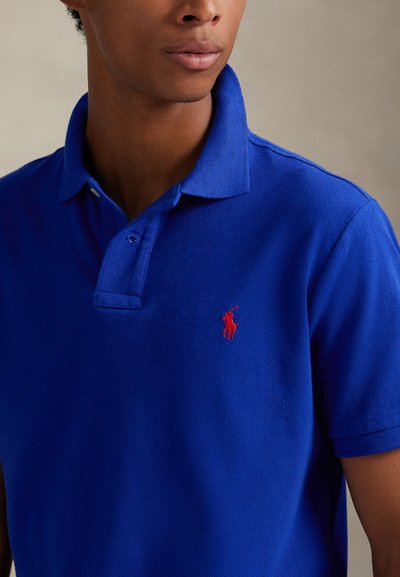 Férfi, aki királykék pólóinget visel, melyen piros hímzett polo játékos logó található a mellkasán, semleges háttér előtt.