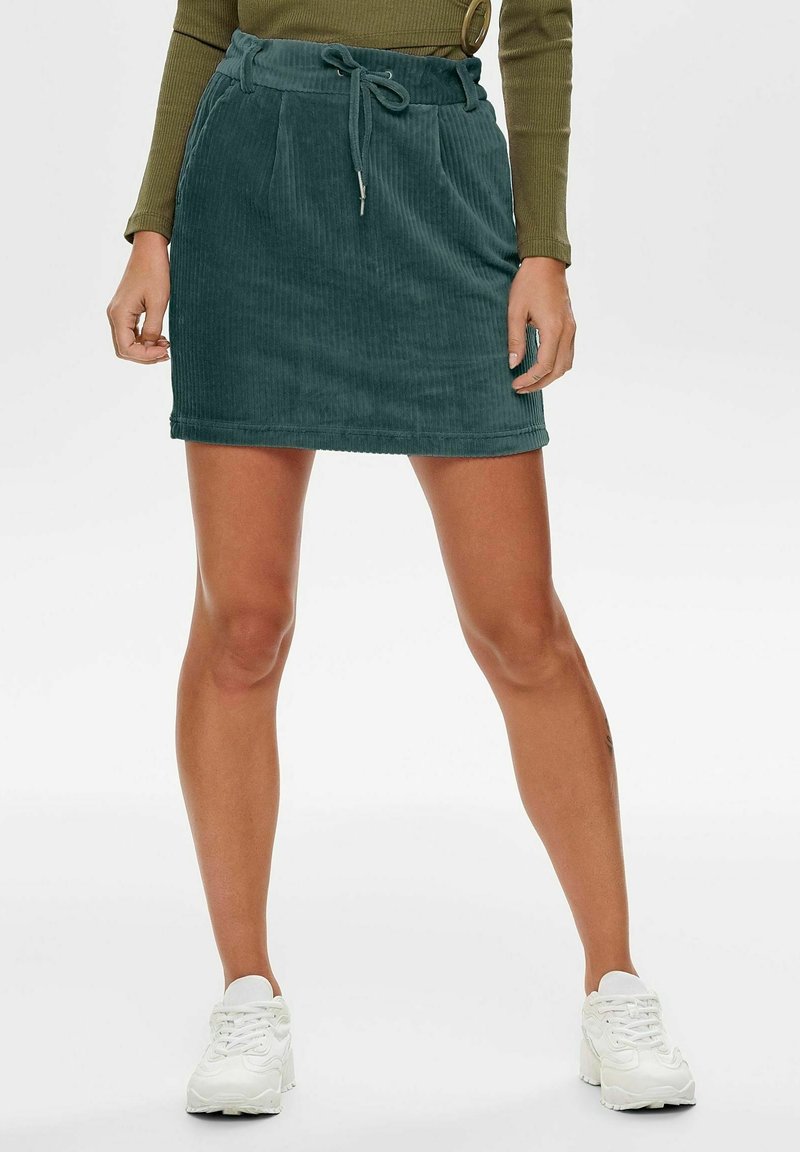 Femme portant une jupe en velours côtelé verte avec une taille à cordon, un haut à manches longues vert olive et des baskets blanches, debout les jambes écartées.