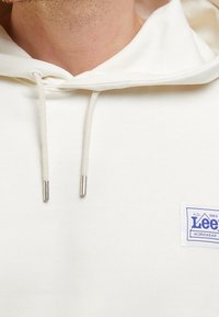 Krémová mikina s kapucí a šňůrkami, s malou obdélníkovou nášivkou loga Lee Workwear na hrudi.
