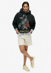 Felpa nera con grafica di uno scorpione e una rosa, pantaloni corti beige e sneaker chiare. La felpa presenta un cappuccio con cordino e una vestibilità oversize.