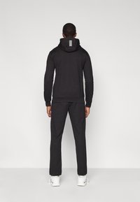 Μαύρο φούτερ με κουκούλα και ταιριαστό παντελόνι από μαλακό ύφασμα. Το φούτερ έχει λογότυπο στην πλάτη με οριζόντιες λωρίδες. Το μοντέλο φοράει λευκά αθλητικά παπούτσια.