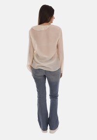 Mujer de espaldas con una blusa transparente color crema de manga larga con cuello con volantes y vaqueros acampanados de talle alto y azul deslavado.