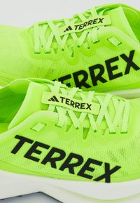 Neon zöld sportcipők fekete "TERREX" márkázással, légáteresztő mesh felsőrésszel, bonyolult fűzéssel és párnázott fehér talppal.