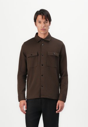 Brown button-up jacket made of textured fabric, featuring a collar, two chest pockets, and black buttons, paired with dark pants.