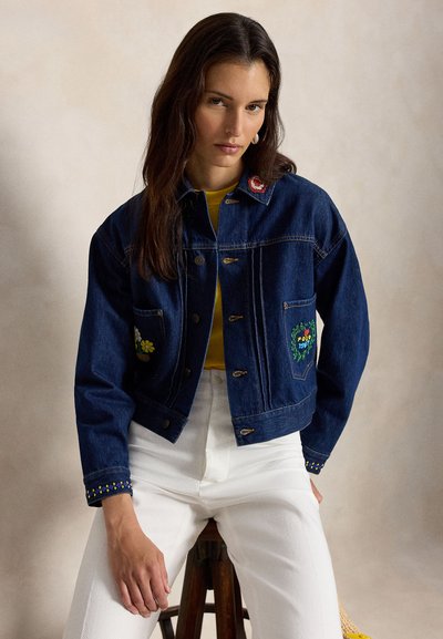 Femeie purtând o jachetă din denim albastru închis cu broderie florală, tricou galben pe dedesubt și pantaloni albi, stând pe un taburet de lemn, având un fundal neutru.