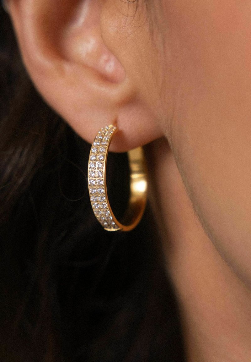 Goldene Creolen mit einer Reihe von klaren Strasssteinen entlang der Vorderkante, glatte Oberfläche und mittlere Dicke. Informelle Trageweise.