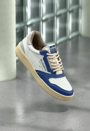 Άσπρο και μπλε sneaker AllSaints με μπεζ σόλα και κορδόνια, αιωρούμενο πάνω από ένα γυαλισμένο σκυρόδεμα δάπεδο με φόντο τοίχο από γυάλινα μπλοκ.