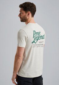 Camiseta de algodón beige claro de manga corta, con texto verde "Pme Legend" y detalles impresos adicionales en la parte posterior.