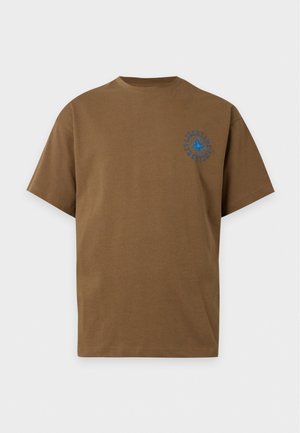 Homme grand à la peau foncée portant une chemise marron à manches courtes par-dessus une chemise noire à manches longues avec des motifs bleus, pantalon foncé et chaussures claires, debout.