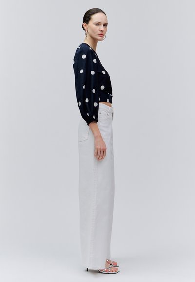 Oldalra néző nő fekete pöttyös blúzban, magas derekú széles fehér nadrágban és ezüst pántos magassarkúban egy egyszerű háttér előtt.