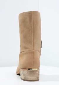 Bottine beige en daim avec un bout arrondi, un petit talon empilé, une fermeture éclair latérale et une garniture dorée au niveau du talon. Texture lisse.