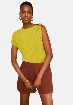 Kvinde iført en ærmeløs gul top med en dekorativ broche på skulderen og højtaljede brune shorts, stående med hænderne foldet.