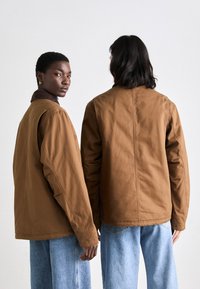 Brune mønstrede jakker med en afslappet pasform, der har en kontrastkrave, båret over lyseblå denimbukser. Bagfra set af to modeller.