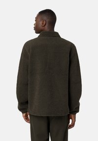 Giacca camicia in fleece verde scuro con superficie testurizzata, colletto e maniche lunghe. Il design presenta una vestibilità comoda e un orlo arrotondato.