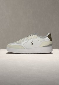 Λευκό sneaker χαμηλού τύπου με μπεζ λεπτομέρειες, σχέδιο με κορδόνια και σκούρο λογότυπο παίκτη πόλο στο πλάι, εκτεθειμένο σε φωτεινή επιφάνεια.