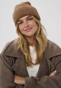Beanie maro cu textură, de tip slouchy, purtat cu o hain�ă supra-dimensionată bej, peste o cămașă albă, evidențiind materiale moi și culori naturale.