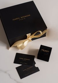 Zwarte Isabel Bernard Parijsdoos met gouden lint, bijpassende fluwelen ringdoos en twee merkkaarten op een lichte ondergrond.