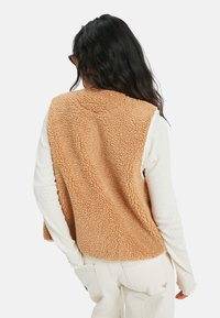 Fleece-Weste in Hellbraun mit einer strukturierten, flauschigen Außenseite. Über einem langärmeligen weißen Oberteil getragen, mit dezenten Stoffdetails.
