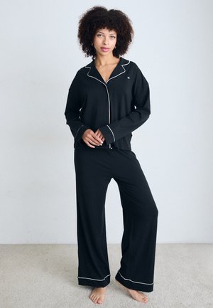 Svart pyjamasset bestående av en knäppbar topp med vit piping och långa ärmar, tillsammans med vida byxor, placerat på en ljus bakgrund.