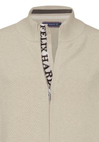 Maglione beige con zip, caratterizzato da una superficie testurizzata, collo alto e zip decorativa con il marchio "FELIX HARDY" in marrone scuro.