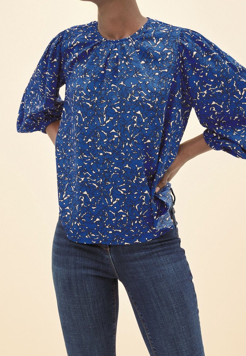 Blusa blu con motivo floreale, realizzata in un tessuto testurizzato, scollatura rotonda e maniche a sbuffo; abbinata a jeans blu scuro.