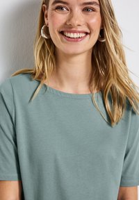 Muntgroene, korte mouwen T-shirt met een brede halslijn. Gladde katoenen stof, minimale stikseldetails, gedragen met zilveren creolen.