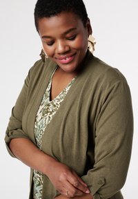 Cardigan vert olive avec des manches retroussées, texture douce et accents de boutons. Assorti à un blouse à motifs dans des tons verts et beiges.